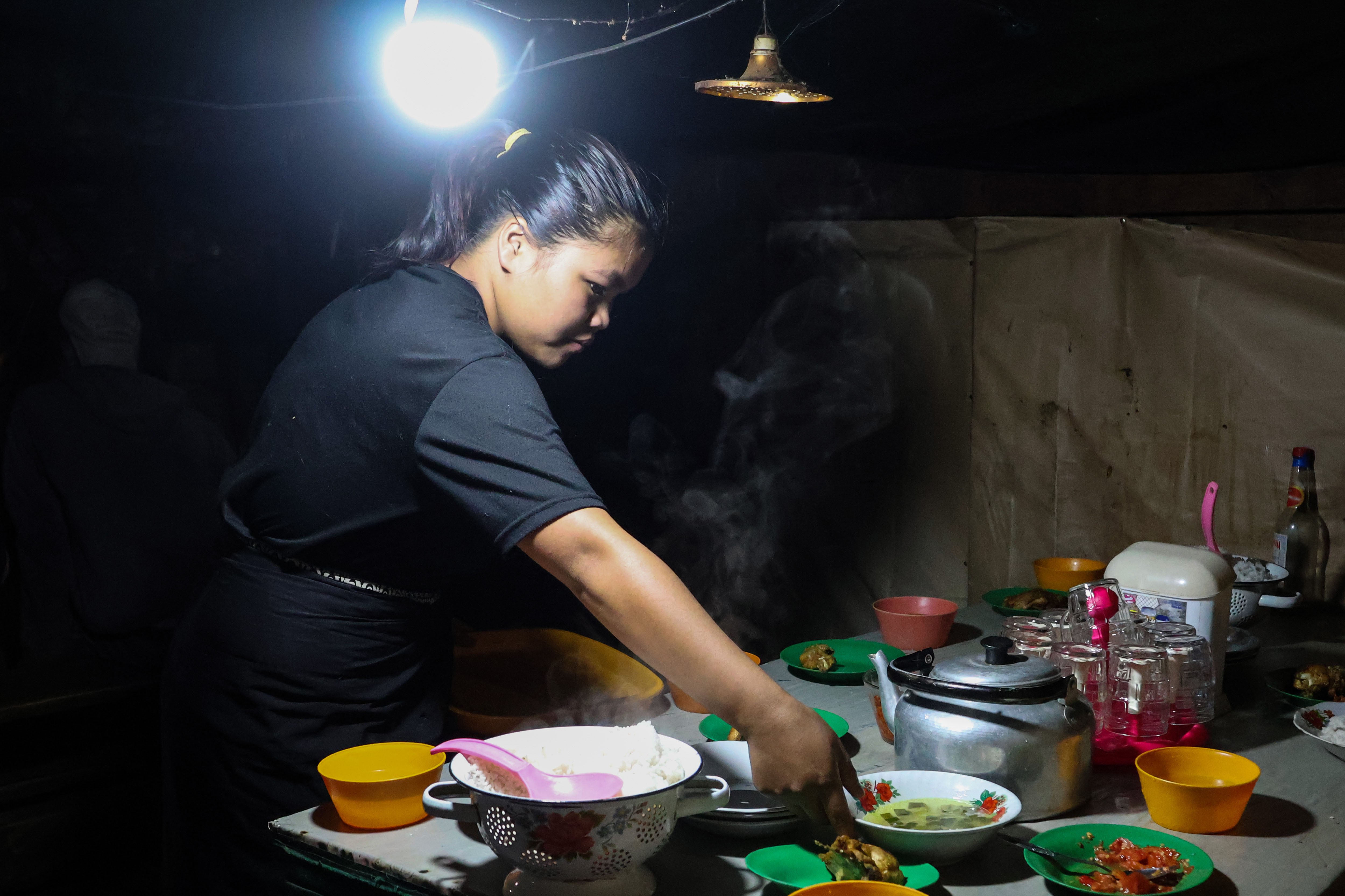 Seorang pelayan menyajikan menu makanan di salah satu warung makan dengan penerangan lampu neon di Desa Embonatana, Kecamatan Seko, Luwu Utara, Sulawesi Selatan.