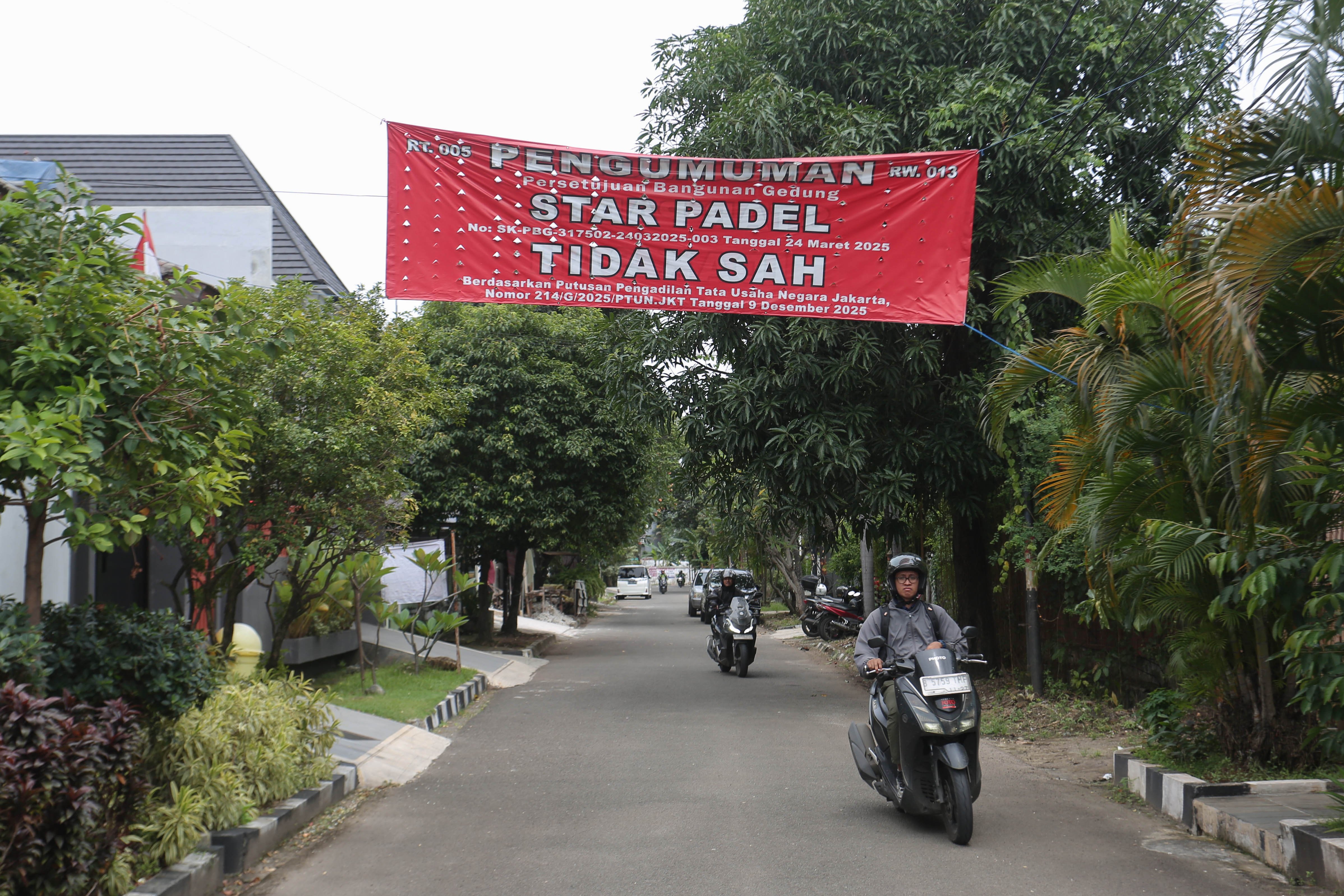 Pengendara sepeda motor melintas di dekat spanduk penolakan lapangan padel di kawasan perumahan, Jalan Pulomas Barat II, Kayu Putih, Pulogadung, Jakarta Timur, Kamis (26/2/2026). Berdasarkan penuturan warga aksi pemasangan spanduk tersebut sebagai bentuk protes terhadap aktivitas olahraga di lapangan padel yang kerap menimbulkan suara bising dan mengganggu ketenangan warga.