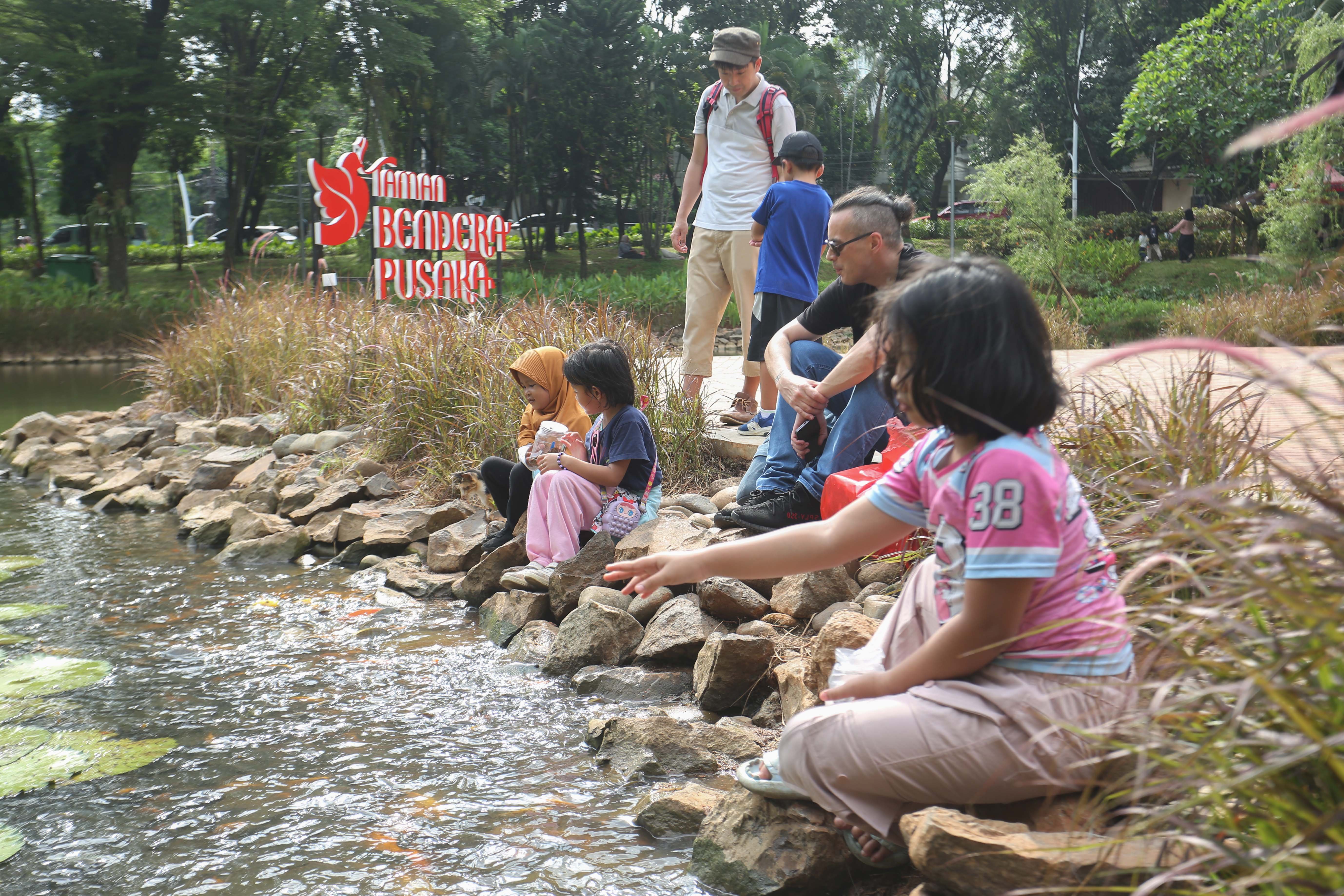 Warga bersama anaknya memberi makan ikan di Taman Bendera Pusaka, Kebayoran Baru, Jakarta Selatan, Sabtu (25/4/2026). Taman seluas 5,6 hektare yang menawarkan suasana asri dan dilengkapi dengan fasilitas seperti area piknik, lapangan olahraga, hingga tempat bermain anak ini menjadi destinasi wisata baru bagi masyarakat dalam menghabiskan waktu akhir pekan.