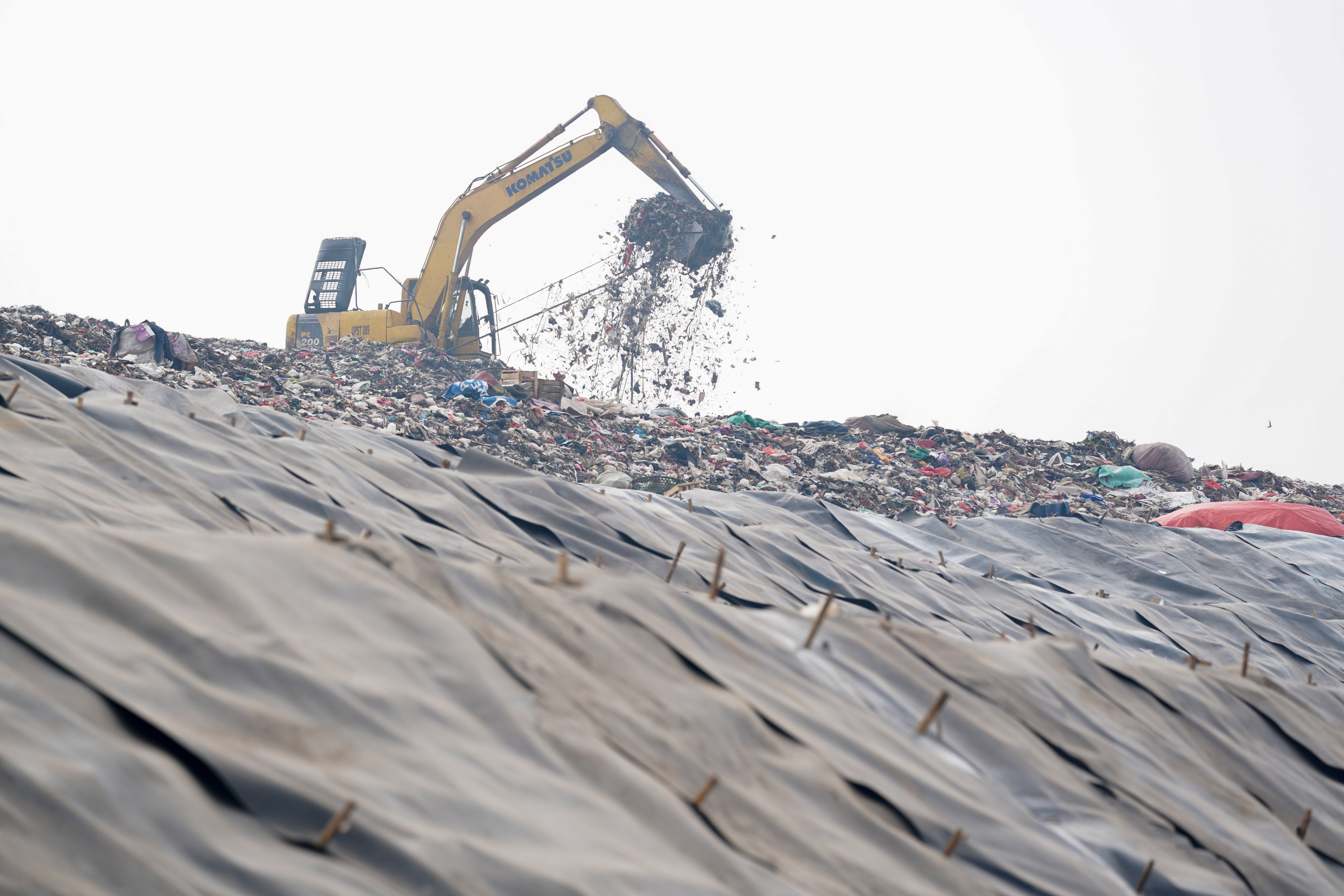 Alat berat eskavator memindahkan sampah di dekat tumpukan sampah yang telah ditutup geomembran di Tempat Pembuangan Sampah Terpadu (TPST) Bantargebang, Kota Bekasi, Jawa Barat, Selasa (4/8/2026). Pemasangan plastik kedap air tersebut merupakan transisi sistem pengolahan sampah dari praktik open dumping menuju controlled landfill yang ditargetkan sepenuhnya diterapkan pada 2029 untuk melindungi area timbunan dan mengurangi cemaran air lindi ke lingkungan.