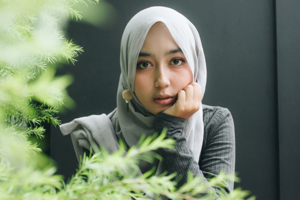 A young woman wearing a hijab is sitting in front of a dark background with green leaves in the foreground, while contemplating on a search query about a prayer to open her aura and charisma.