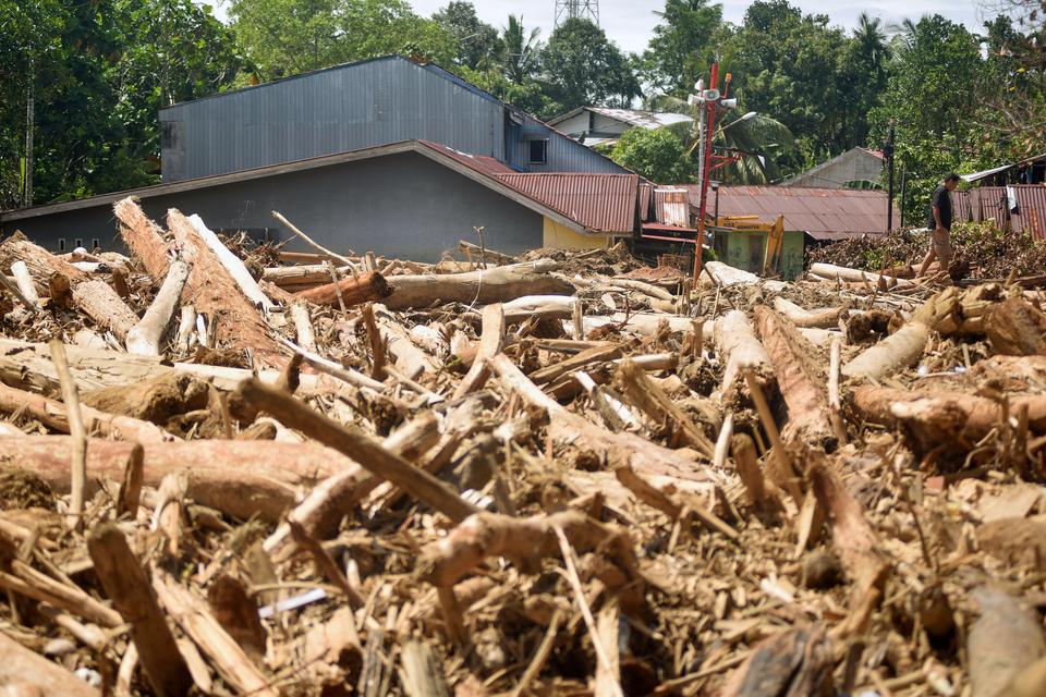 Warga melintas di antara tumpukan gelondongan kayu di permukiman di Tabiang Bandang Gadang, Nanggalo, Padang, Sumatera Barat, Selasa (9/12/2025). Gelondongan kayu yang terbawa banjir bandang sejak dua pekan lalu masih tersangkut di wilayah itu.