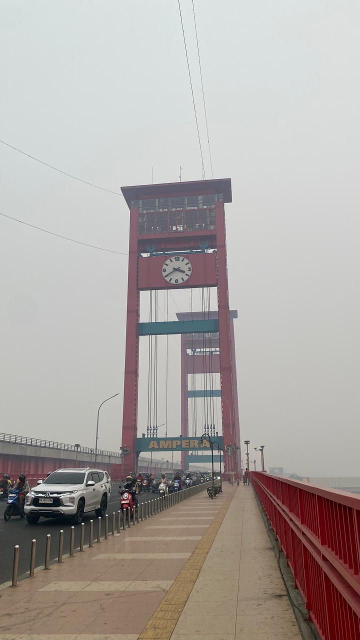 Menara Jembatan Ampera diselimuti kabut asap; kendaraan dan pejalan kaki melintas di atas jembatan.